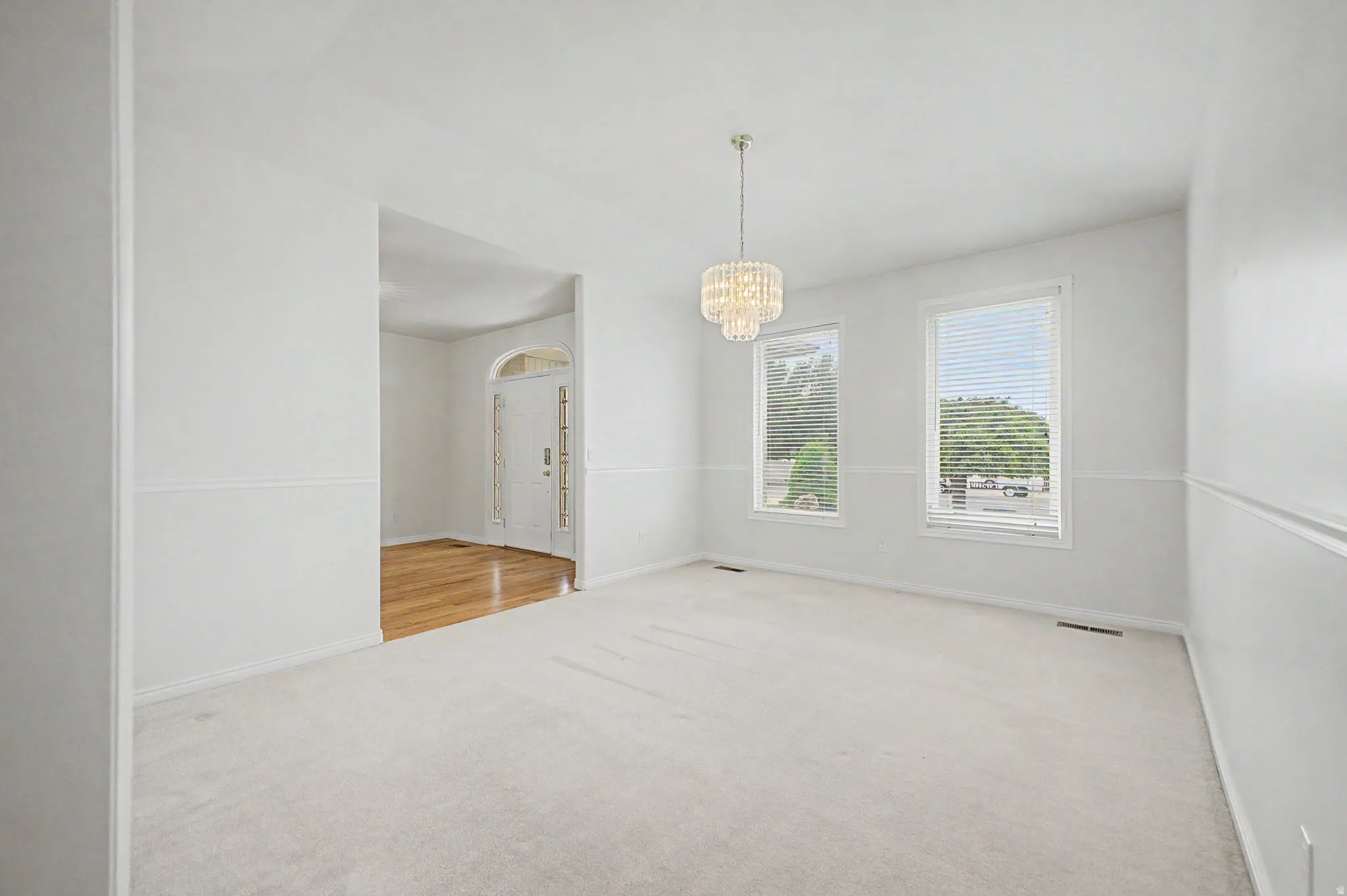 Carpeted room featuring a tiered crystal chandelier, two windows with blinds, and white wall paneling