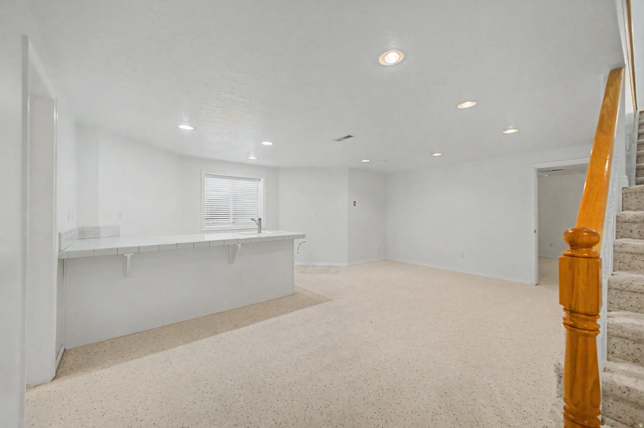 Spacious room featuring a bar with a sink and countertop, recessed lighting, light-colored carpet flooring, a window with blinds, and a wooden handrail beside carpeted stairs