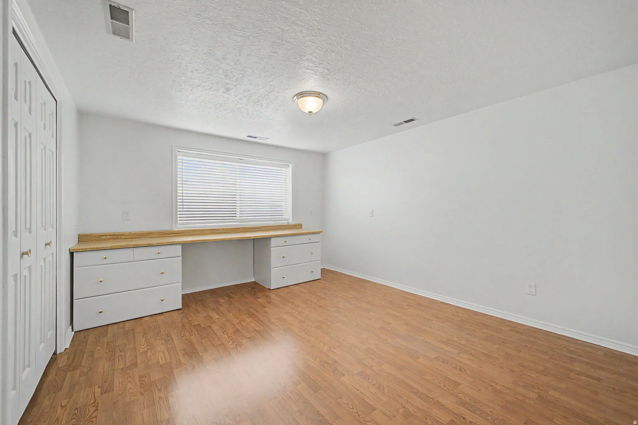 Room featuring wood-finish flooring, a built-in desk with a light wood-finish countertop, white cabinetry with gold-tone hardware, and a window with horizontal blinds