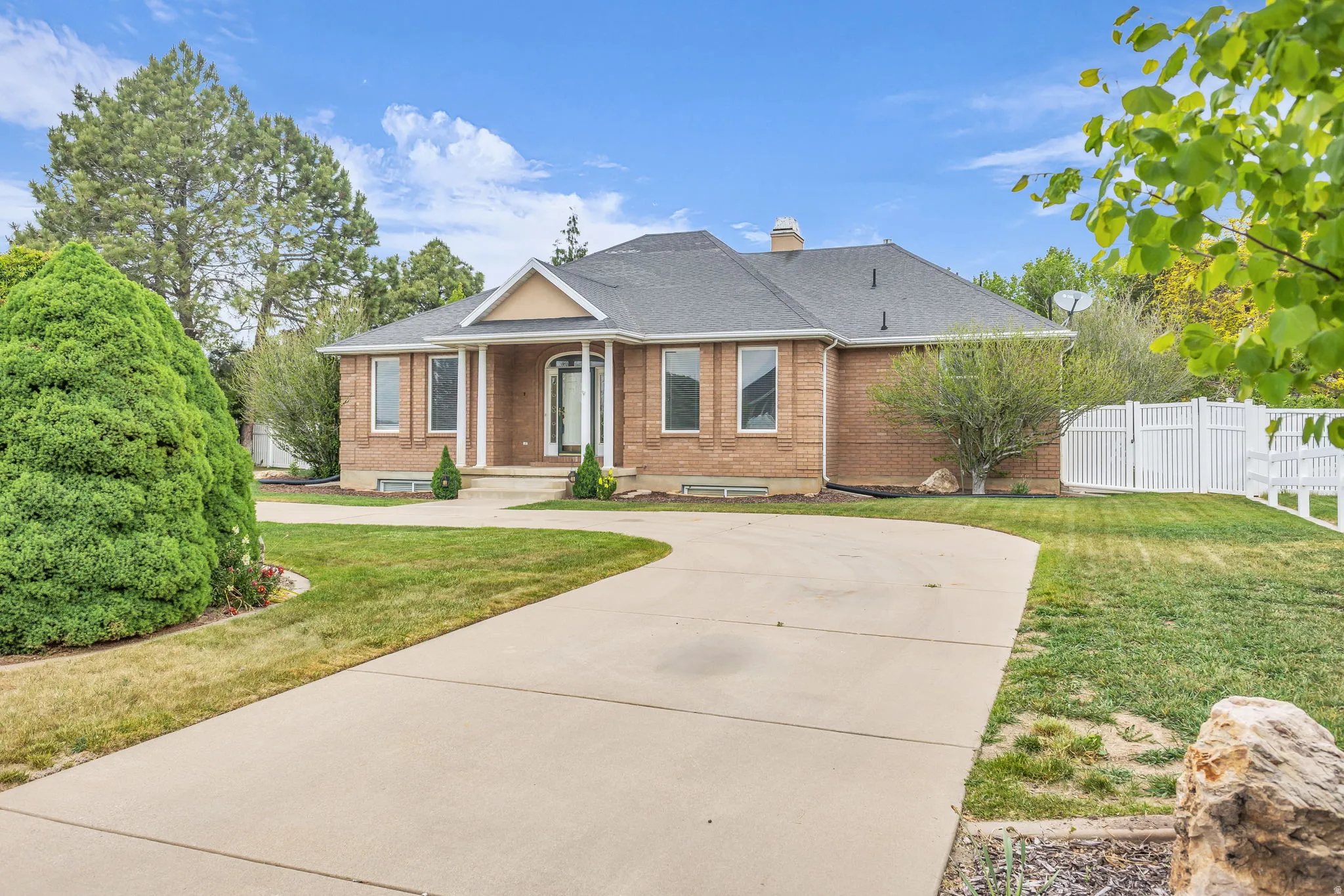 Brick exterior with a prominent portico entry, large front windows, a dark shingle roof, a concrete driveway, and a white fence