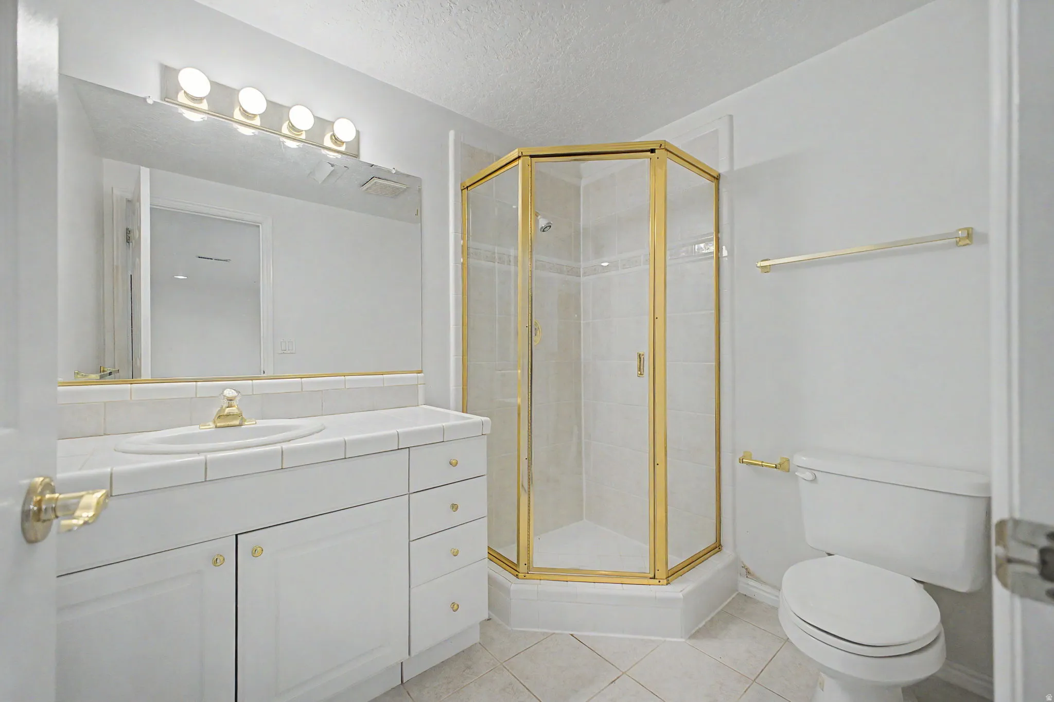 Bathroom featuring a corner shower with a gold-finish frame, a vanity with a tiled countertop and integrated sink, white cabinetry with gold-tone hardware, and a large wall-mounted mirror with overhead lighting