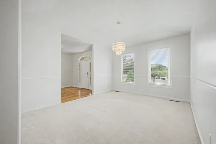Carpeted room featuring a tiered crystal chandelier, two windows with blinds, and white wall paneling