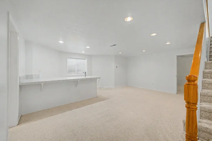 Spacious room featuring a bar with a sink and countertop, recessed lighting, light-colored carpet flooring, a window with blinds, and a wooden handrail beside carpeted stairs