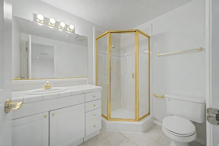 Bathroom featuring a corner shower with a gold-finish frame, a vanity with a tiled countertop and integrated sink, white cabinetry with gold-tone hardware, and a large wall-mounted mirror with overhead lighting