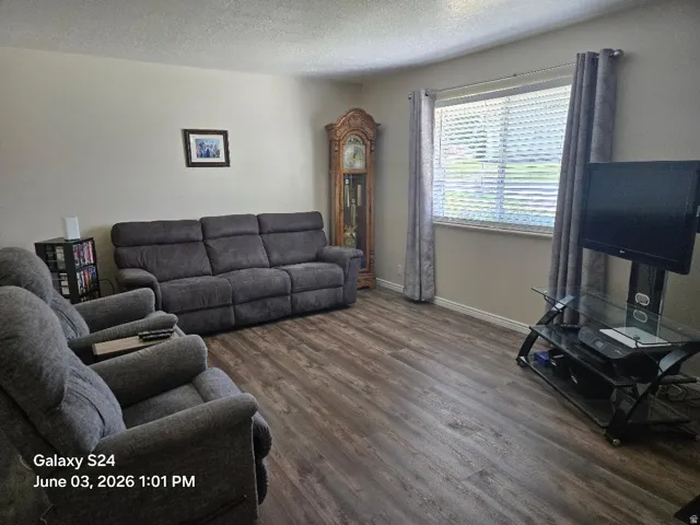 Living area featuring wood-finish flooring, neutral wall tones, a large window with blinds, and ceiling trim
