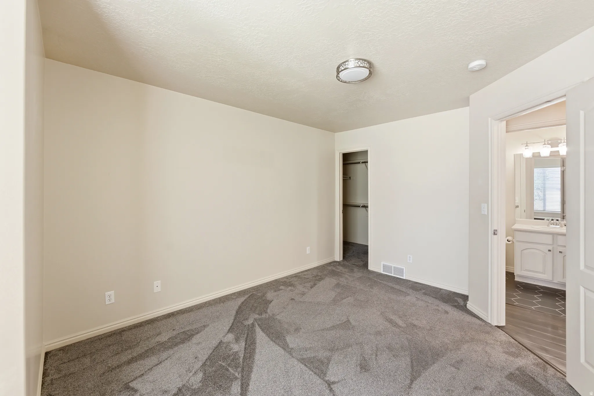 Carpeted Primary Bedroom featuring a ceiling-mounted light fixture, an open closet with built-in shelving, and a doorway revealing a bathroom with a white vanity and patterned flooring