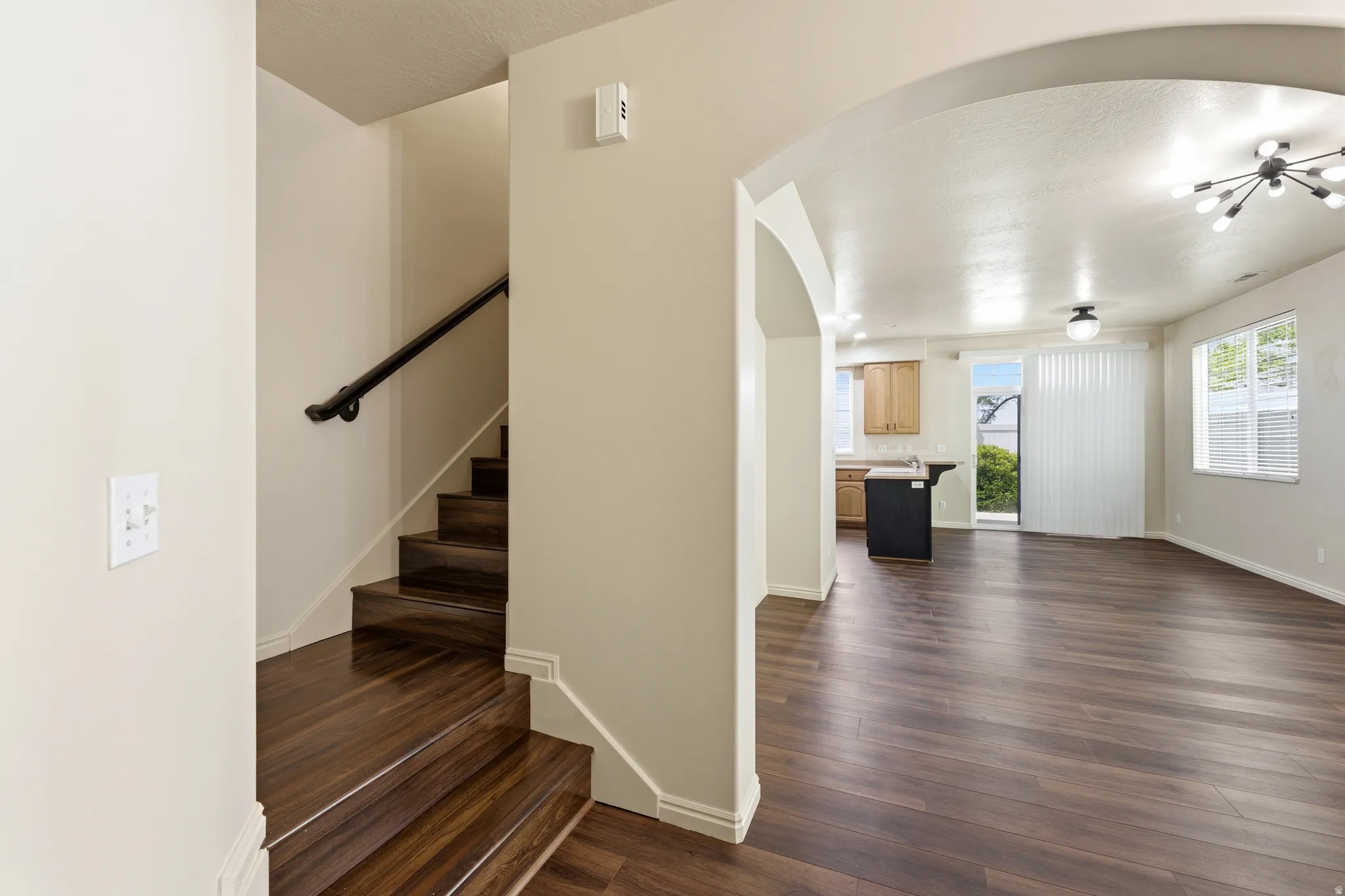 Wood-finish flooring throughout, featuring an arched entryway, a staircase with a dark wood handrail, a kitchen area with light wood cabinetry, and a sliding glass door