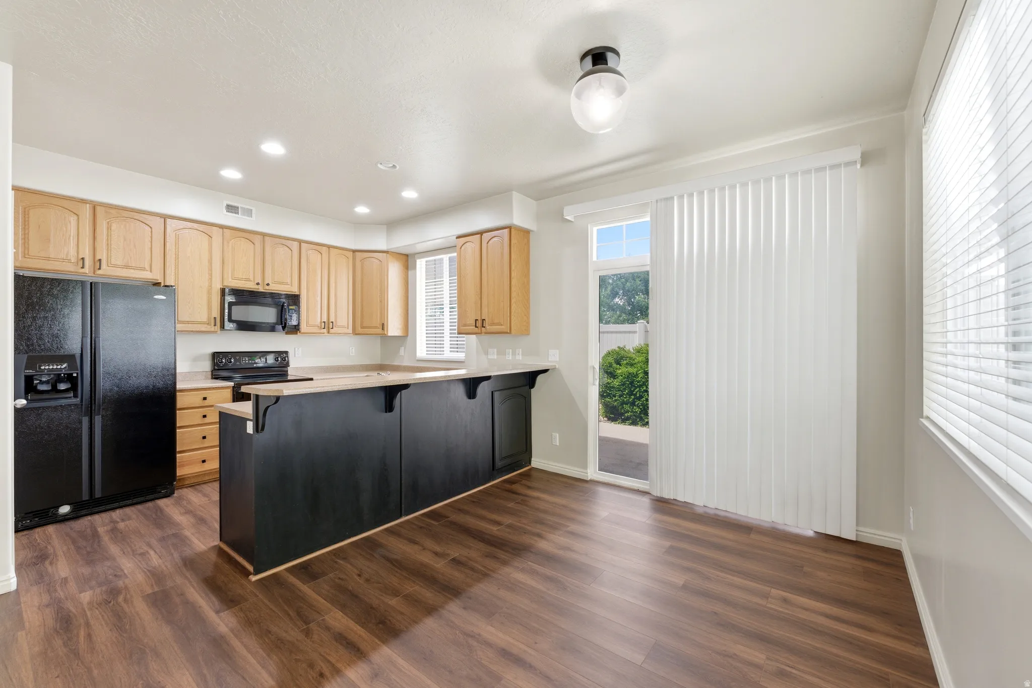 Kitchen featuring wood-finish flooring, light wood cabinetry, black appliances, a central island with counter seating, and a sliding glass door