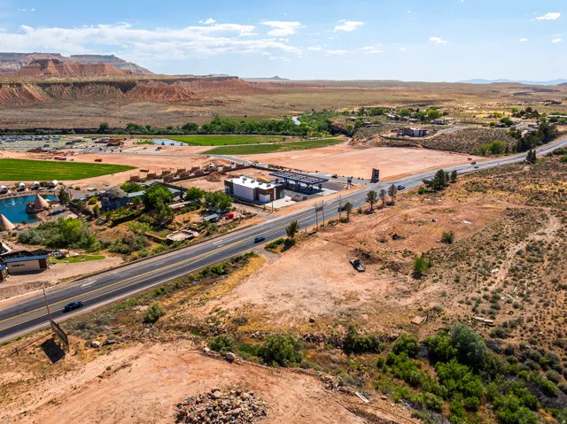Expansive desert landscape featuring a paved road, scattered buildings, and distant red rock formations under a clear sky