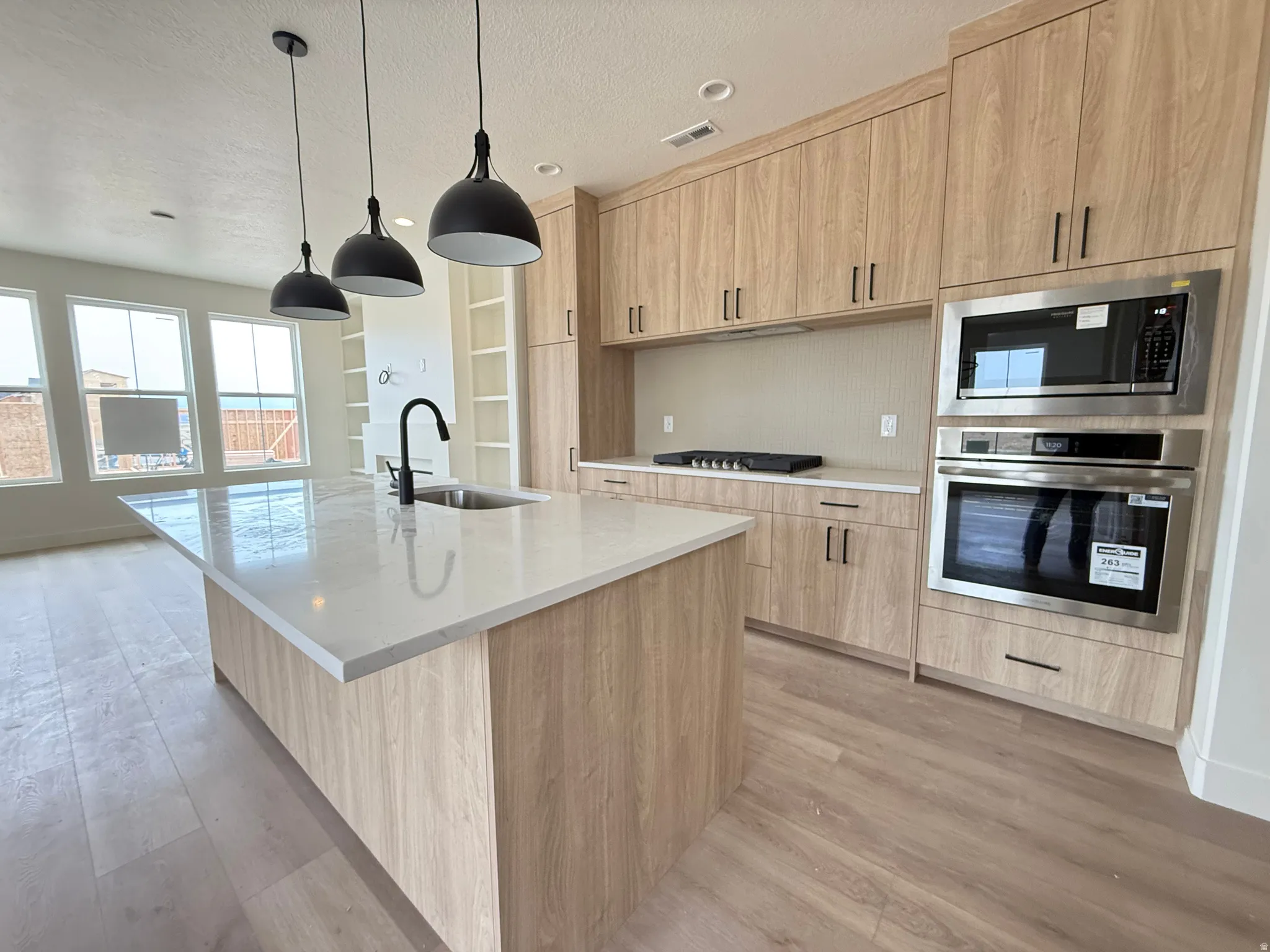 Modern kitchen featuring a large island with a surface-mounted sink, three pendant lights, wood-finish cabinetry, built-in stainless steel appliances, and a gas cooktop