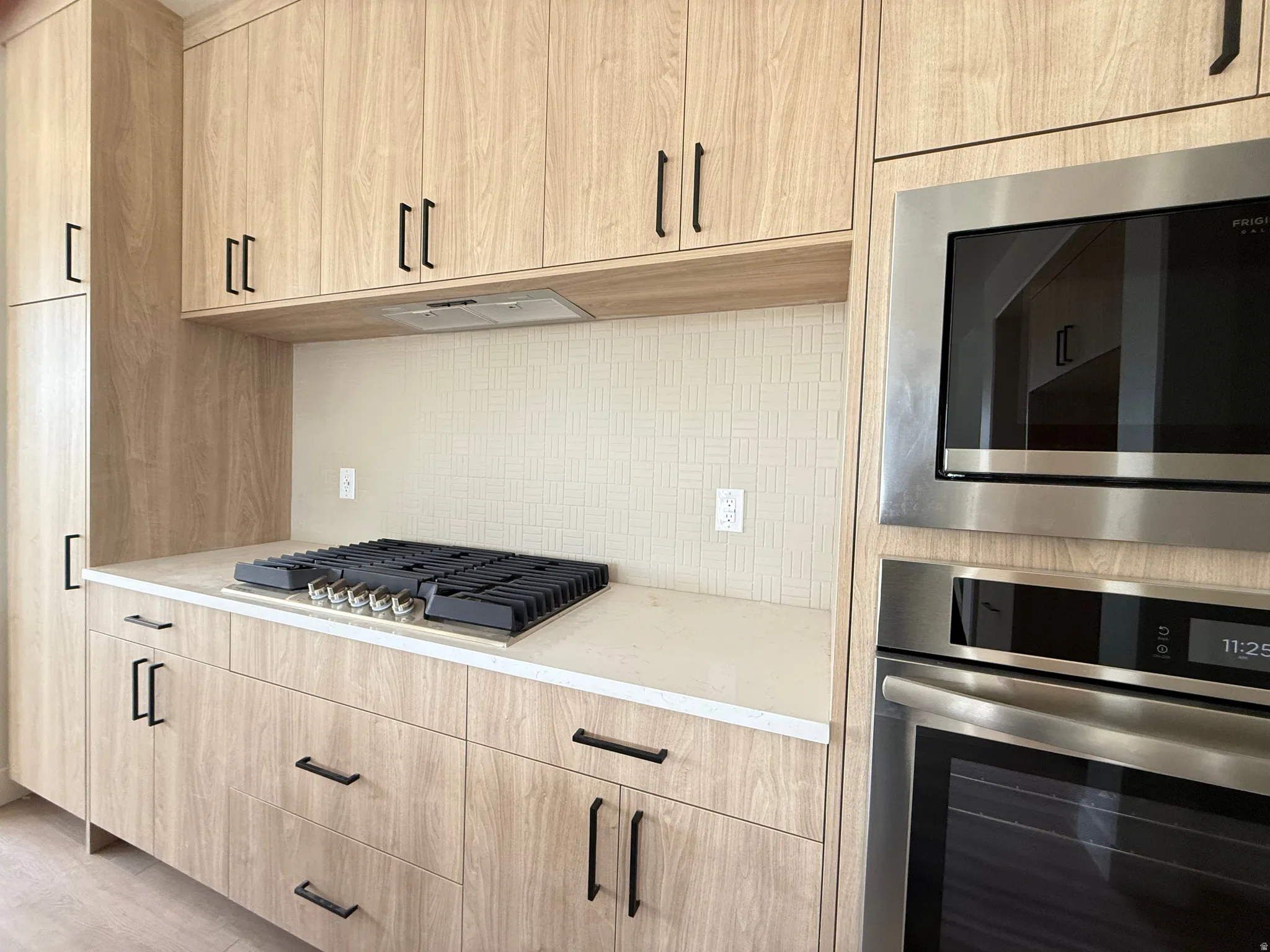 Contemporary kitchen featuring wood-finish cabinetry with matte black hardware, integrated gas cooktop, textured tile backsplash, and built-in stainless steel microwave and oven