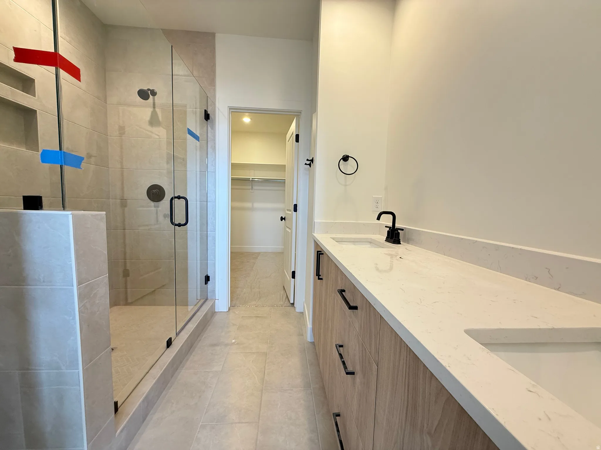 Contemporary bathroom featuring a spacious glass-enclosed shower with tile surround and integrated shelving, dual vanity with undermount sinks, light-toned countertops, and wood-finish cabinetry with matte black hardware