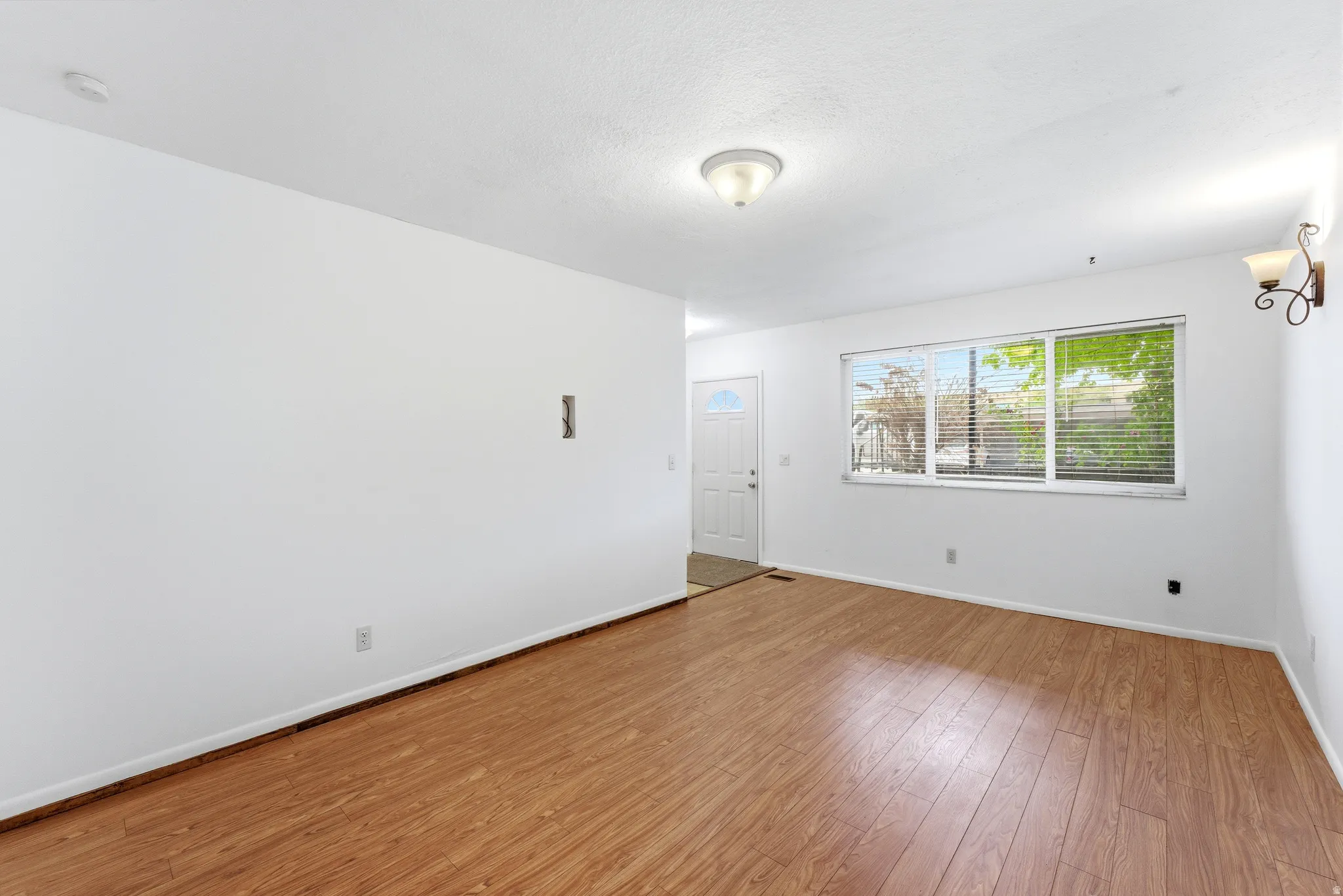 Expansive interior space featuring wood-finish flooring, a large window with horizontal blinds, and a white entry door with an arched glass insert