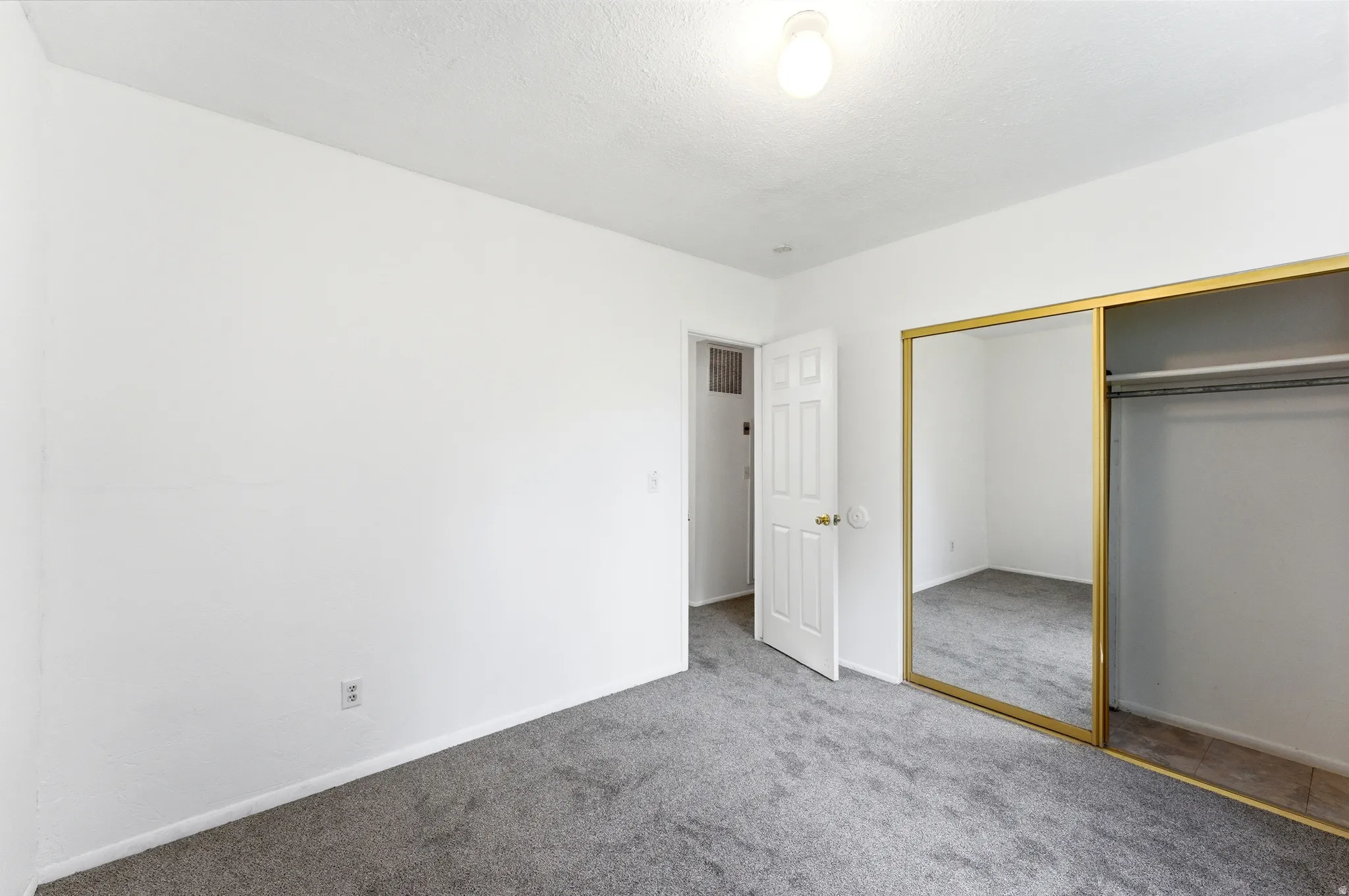 Bedroom featuring gray carpet, white painted walls, a standard white door, a ceiling-mounted light fixture, and a mirrored closet with gold-tone framing