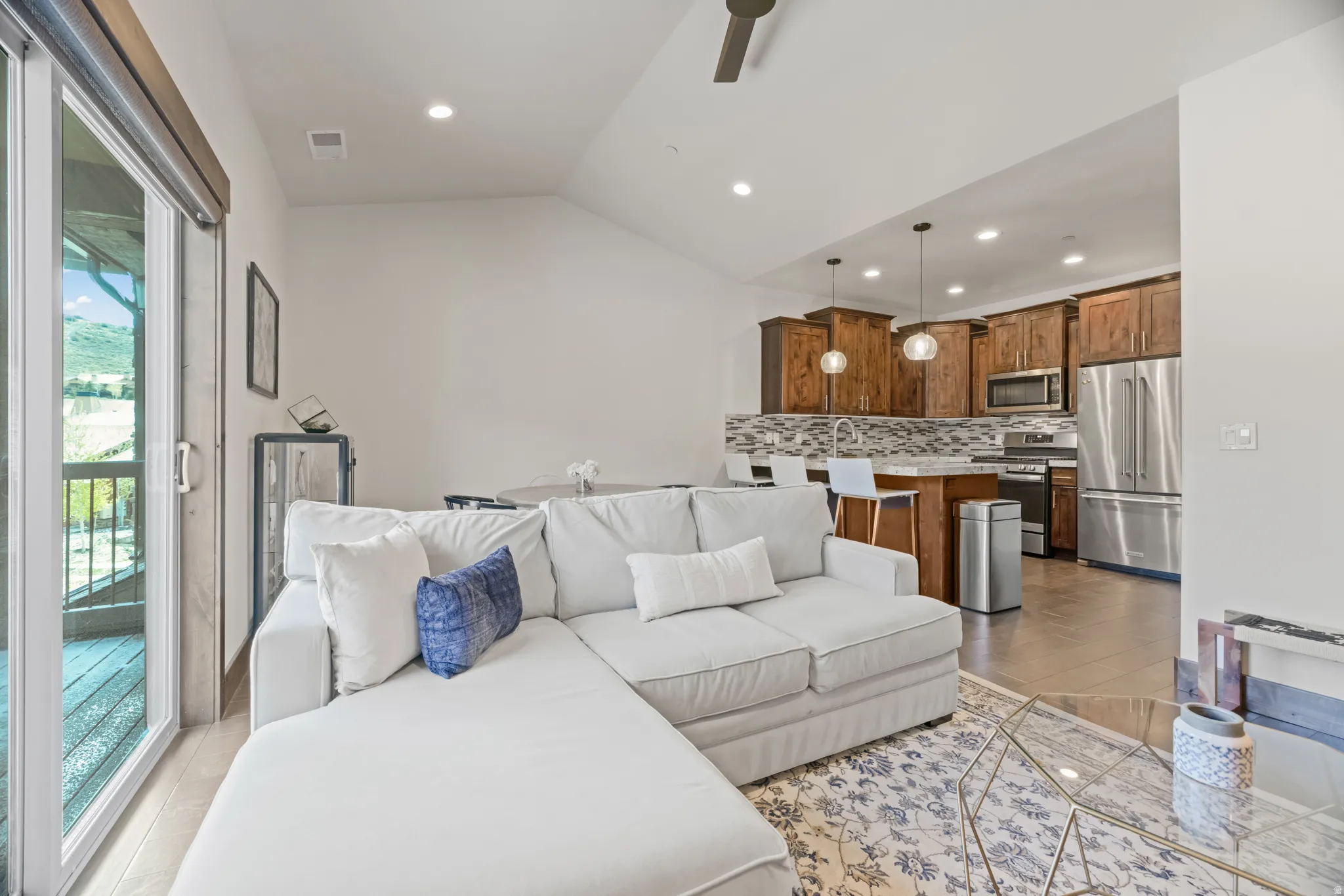 Open-concept living space featuring wood-finish cabinetry, stainless steel appliances, a multi-tone tile backsplash, recessed lighting, and a sliding glass door providing access to an outdoor area