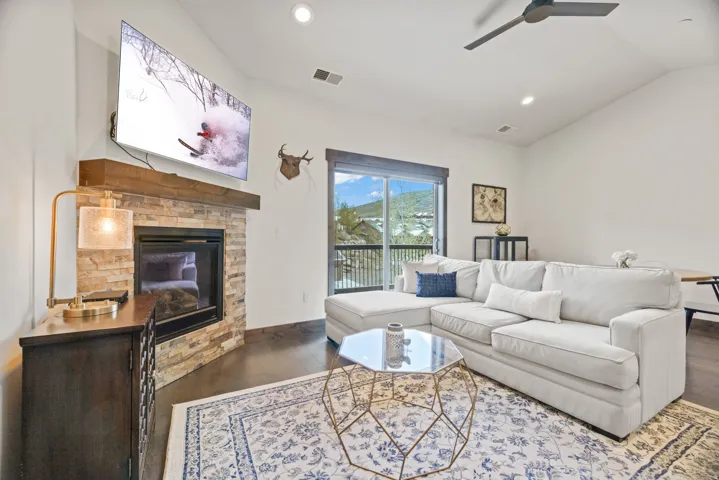 Living room featuring a stacked stone fireplace with a wood mantle, an attached flat-screen television, dark wood-finish flooring, and a sliding glass door opening to an outdoor balcony