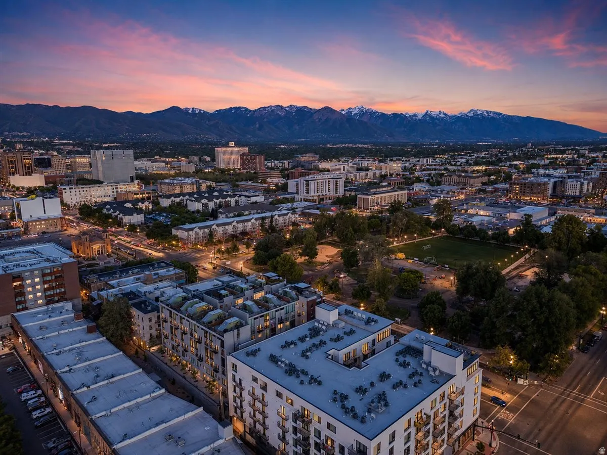 Cityscape featuring a prominent mountain range, urban park space, numerous residential and commercial buildings, illuminated city streets, and a vibrant sunset backdrop