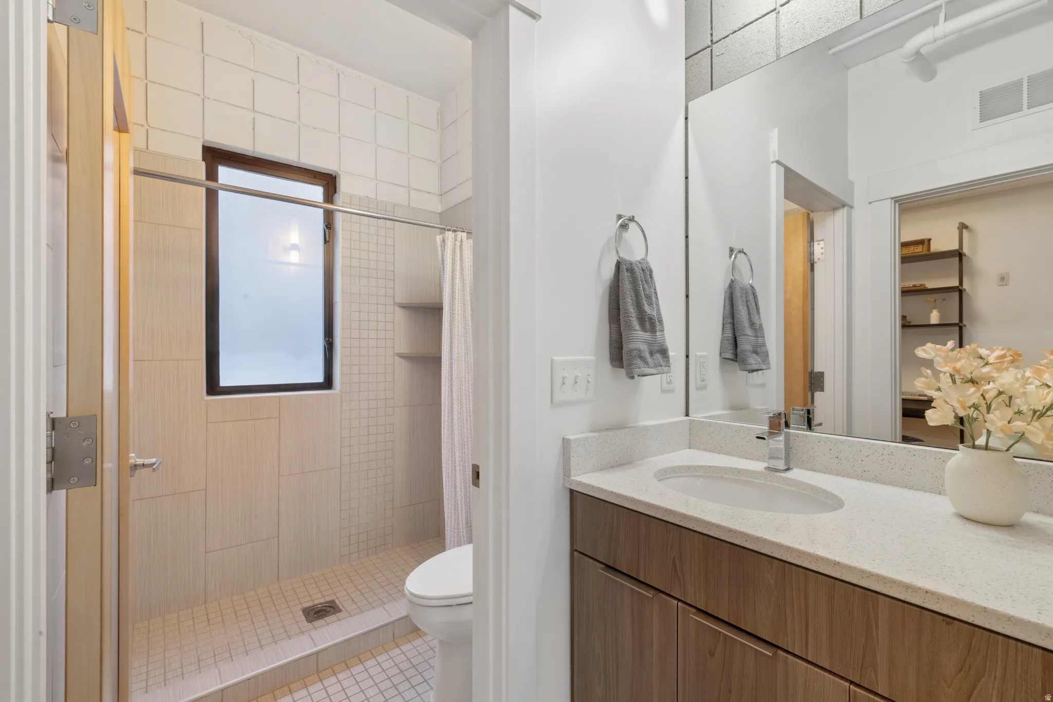 Bathroom featuring a wood-finish vanity with a light-colored countertop and an integrated sink, a large wall-mounted mirror, a tiled shower with a window and built-in shelving, and a toilet