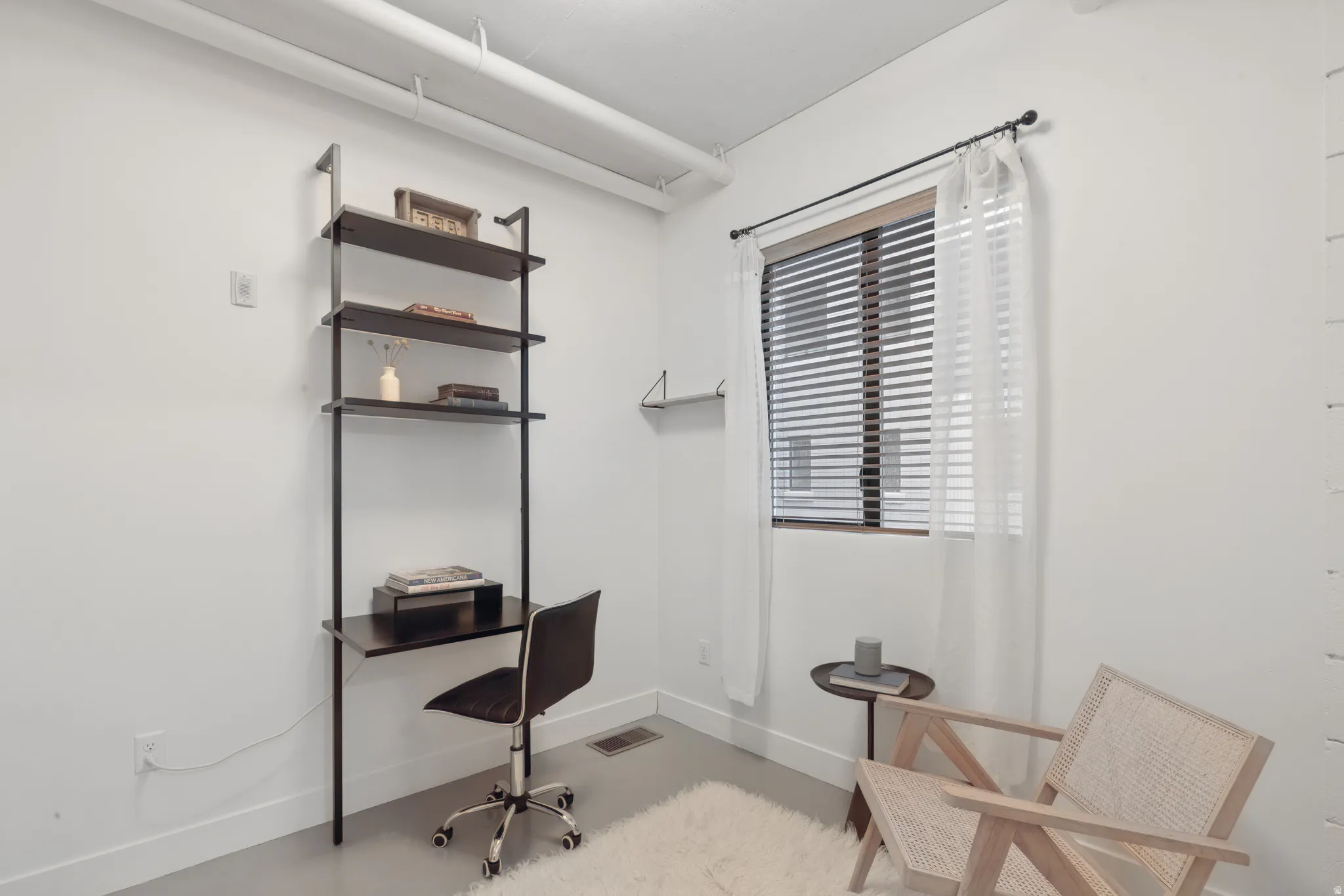 Bright room featuring a window with horizontal blinds and sheer drapes, a built-in wall unit with shelving and an integrated desk, and light-colored flooring