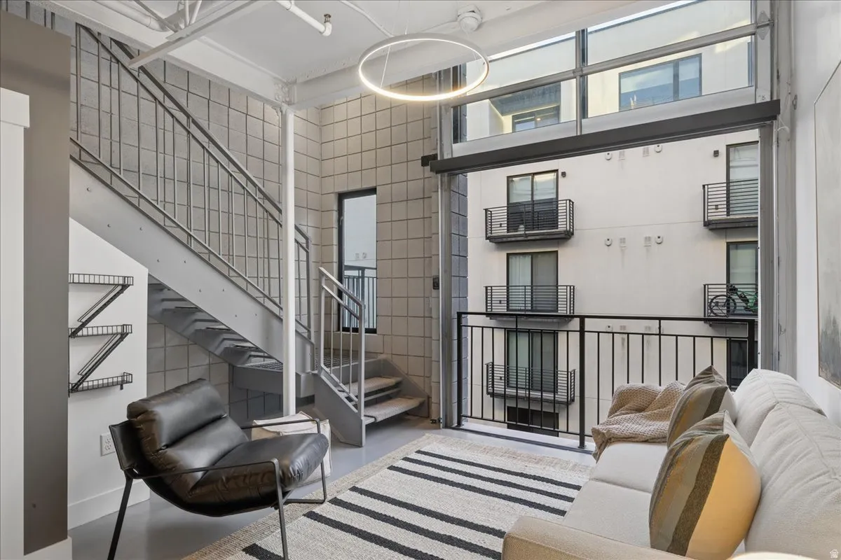 Open-concept living space featuring high ceilings with exposed ductwork, industrial-style staircase with metal railings, full-height window wall with balcony access, and polished concrete flooring
