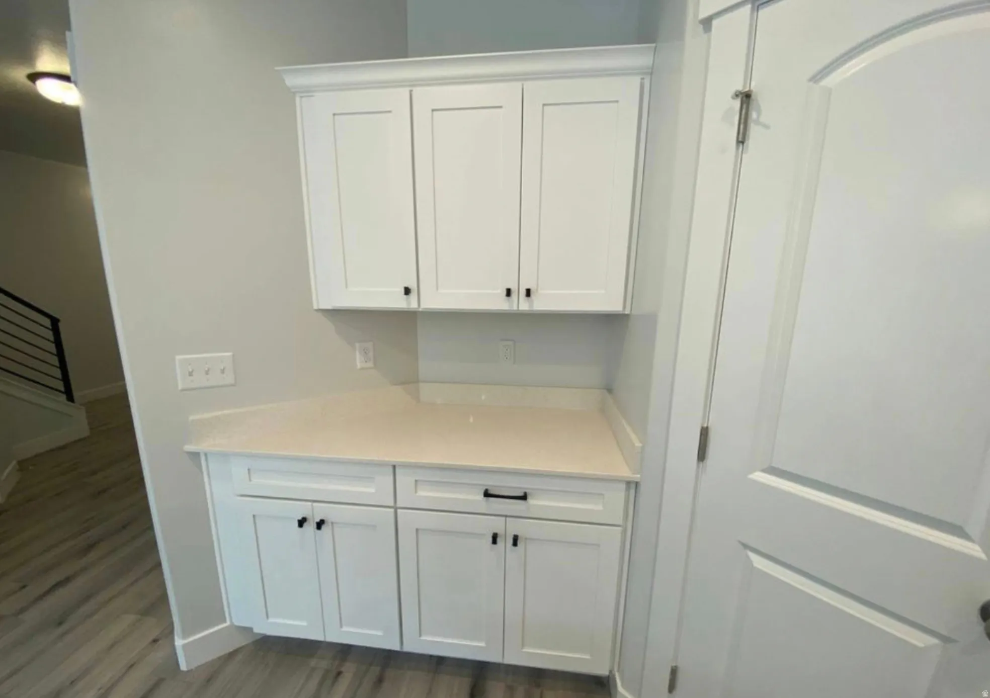 White shaker-style cabinetry with matte black hardware, light-toned countertop, light gray walls, wood-finish flooring, and a white interior door