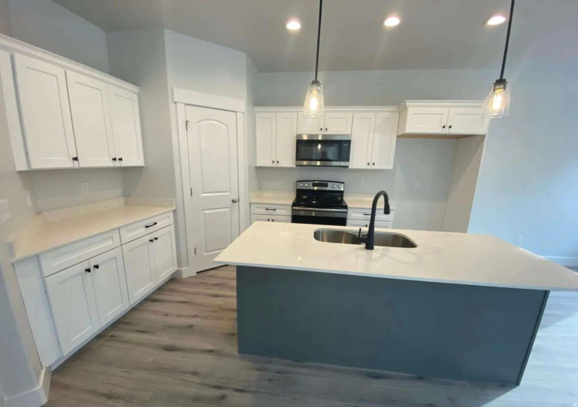Contemporary kitchen featuring white cabinetry, light-toned countertops, a central island with an integrated sink and matte black faucet, wood-finish flooring, and pendant lighting
