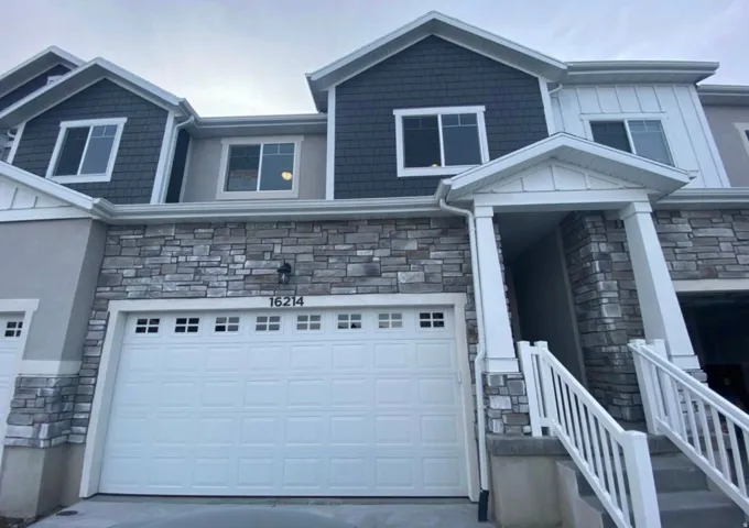 Modern townhouse exterior featuring stone veneer siding, decorative shake siding, multiple windows, a panel garage door with decorative lites, and a covered entry with white railings