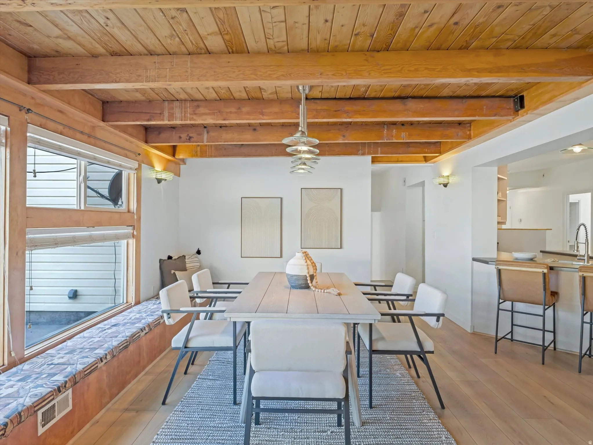 Dining area featuring a wood-planked ceiling with exposed beams, wood-finish flooring, contemporary chandeliers, a window seat with cushioned top, and a pass-through opening to the kitchen