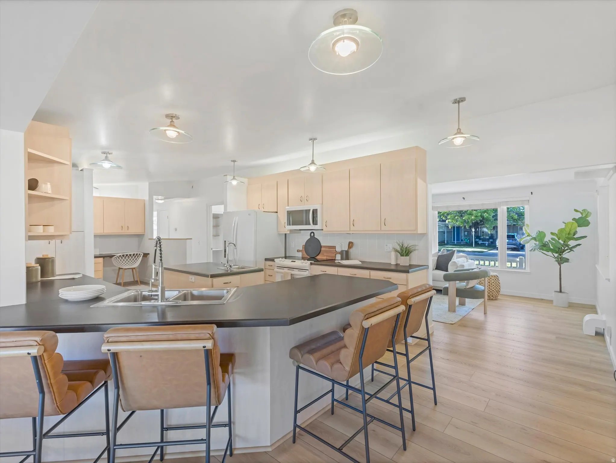 Kitchen featuring a peninsula island with an undermount double sink, light wood-finish cabinetry, dark countertops, white tile backsplash, and light wood-finish flooring