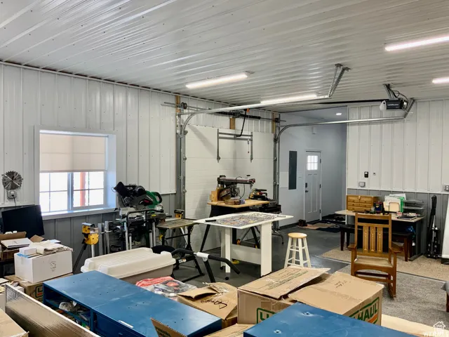 Spacious utility room featuring white corrugated wall panels, a large window, fluorescent overhead lighting, and an overhead garage door