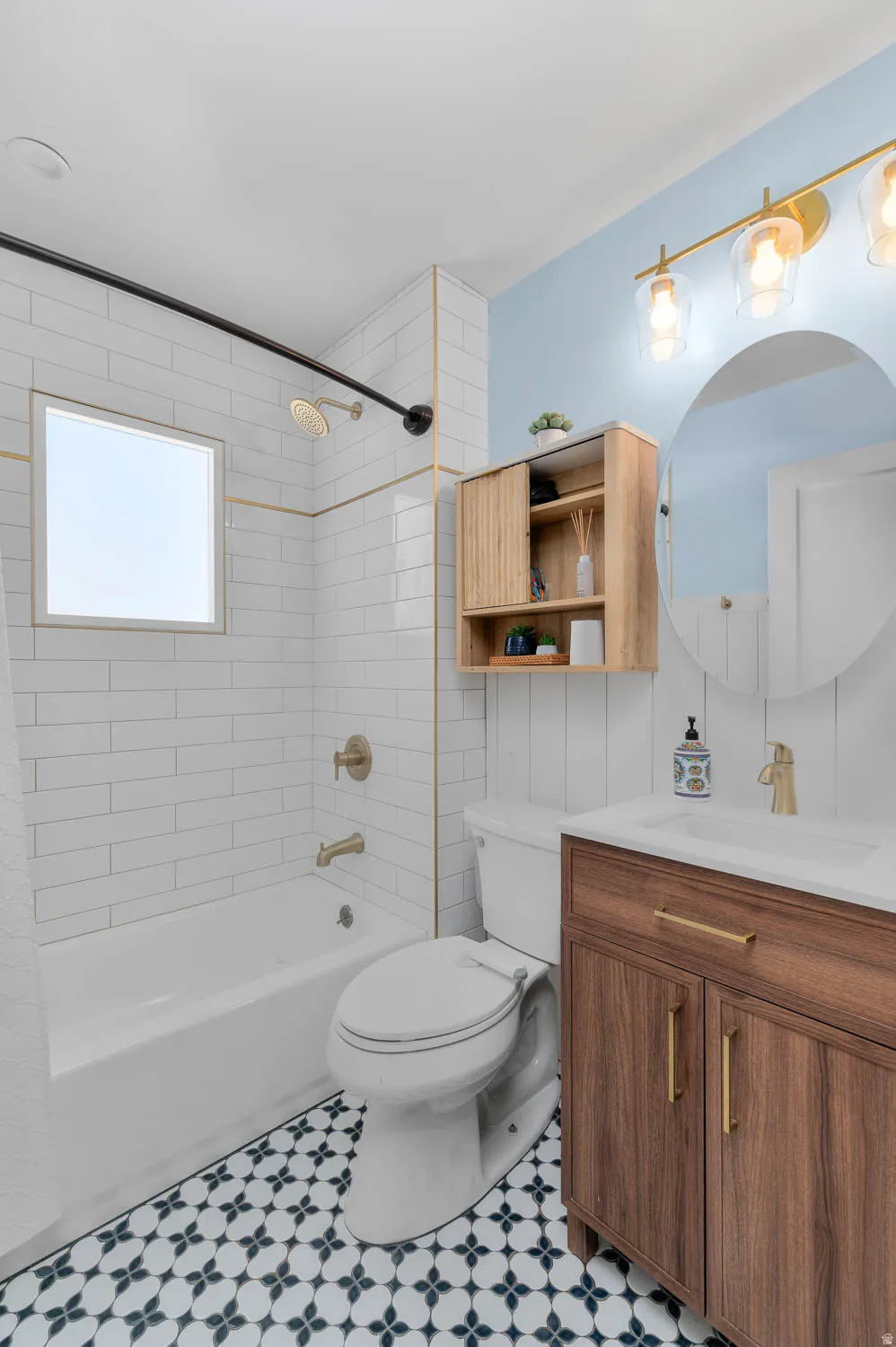 Bathroom featuring patterned floor tiling, a white subway tile tub surround with gold-finish trim, a wood-finish vanity with gold-tone hardware, a circular mirror, and a three-light vanity fixture