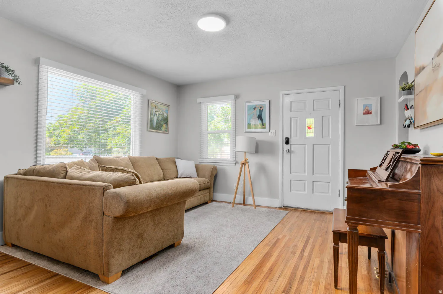 Spacious living area featuring light wood-finish flooring, two large windows with blinds, and a paneled entry door with a decorative glass insert