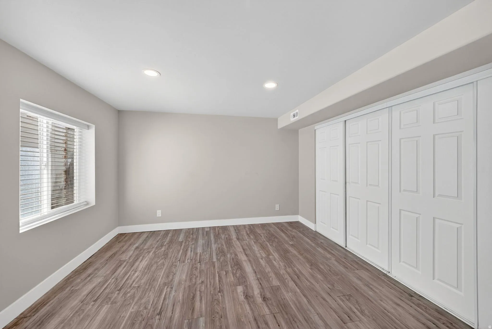 Spacious room featuring wood-finish flooring, neutral-toned walls, a large window with blinds, recessed lighting, and expansive bi-fold closet doors