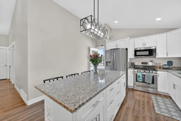 Kitchen featuring a central island with a speckled countertop, white cabinetry, stainless steel appliances, wood-finish flooring, and a modern geometric light fixture