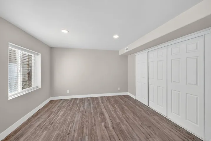Spacious room featuring wood-finish flooring, neutral-toned walls, a large window with blinds, recessed lighting, and expansive bi-fold closet doors