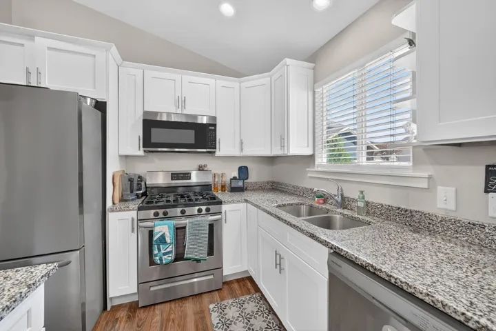 Kitchen featuring white shaker-style cabinetry, stainless steel appliances, speckled stone countertops, a double basin sink, and wood-finish flooring