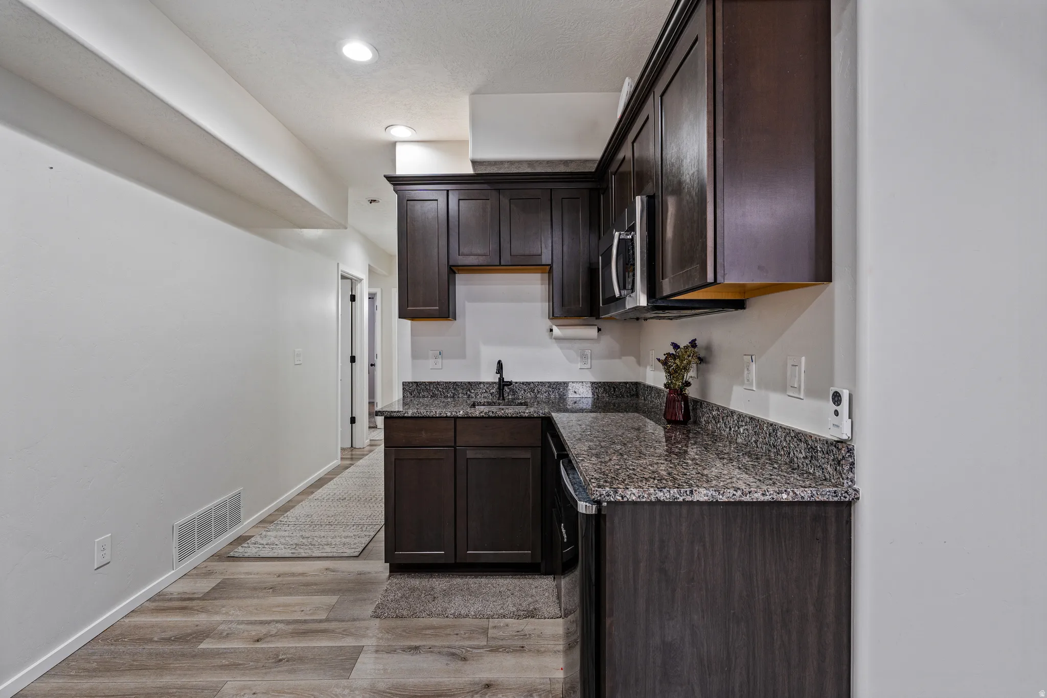 Kitchenette featuring dark wood cabinetry, speckled countertops, stainless steel microwave, and wood-finish flooring
