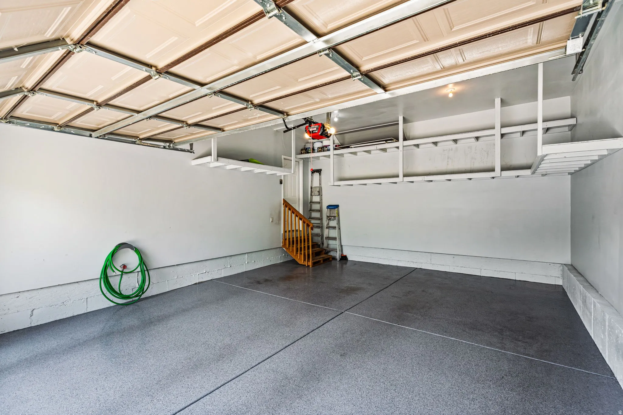 Spacious garage featuring a durable epoxy-coated floor, overhead door, built-in shelving, and a wooden staircase