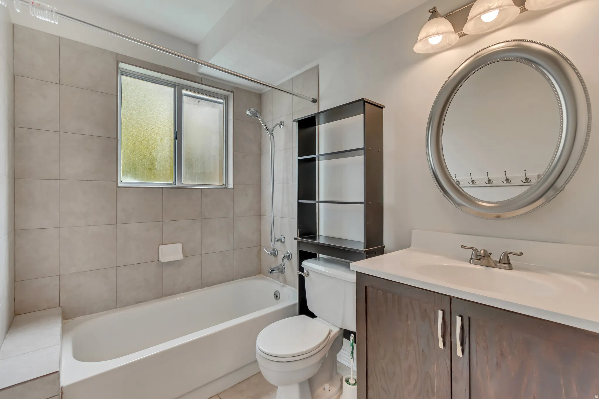 Bathroom featuring a single vanity with a wood-finish cabinet and white countertop, an oval mirror, a three-light fixture, a tub/shower combination with light-toned wall tiling, and a window with obscured glass