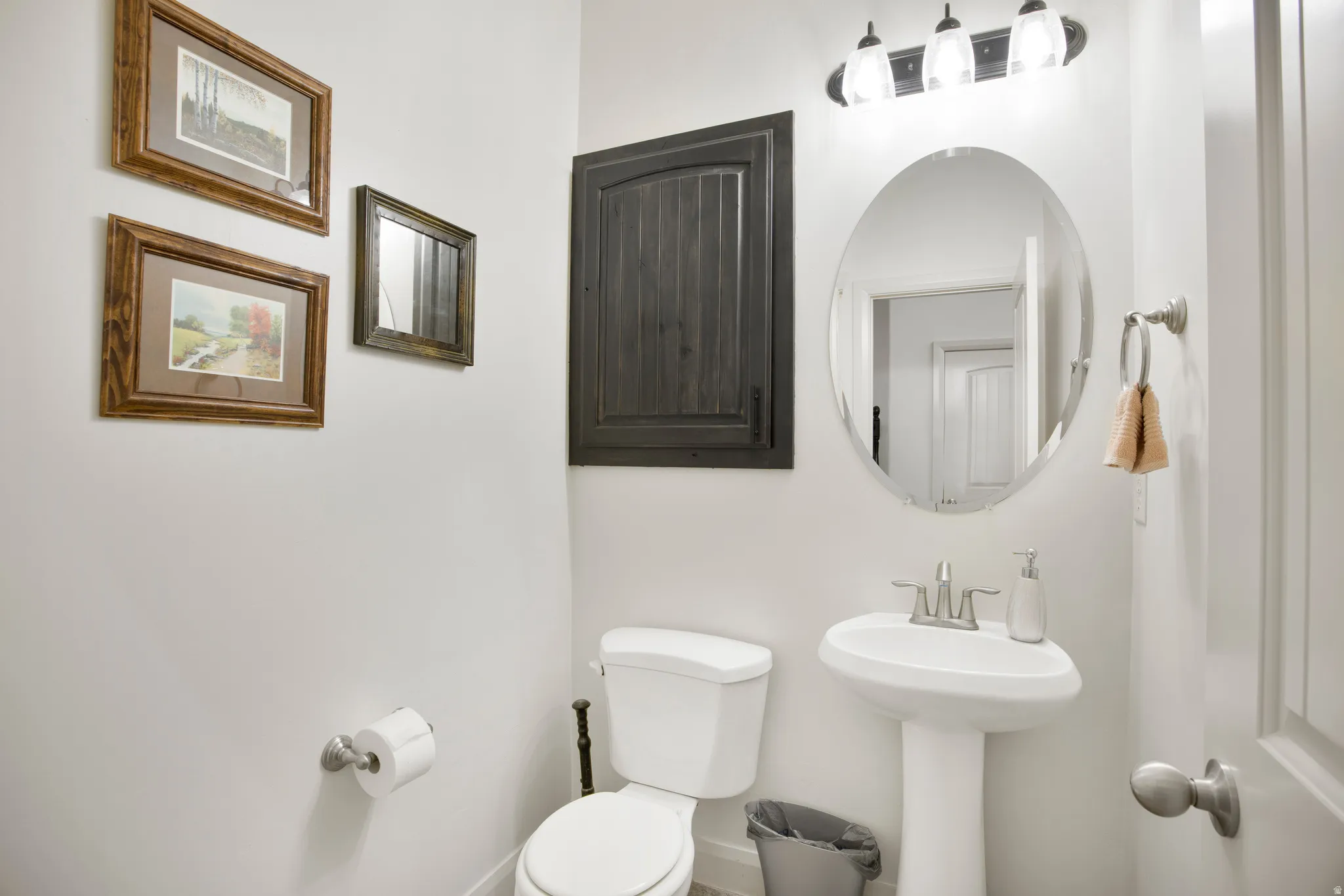 White pedestal sink with brushed nickel faucet, illuminated by a three-light vanity fixture, complemented by an oval mirror
