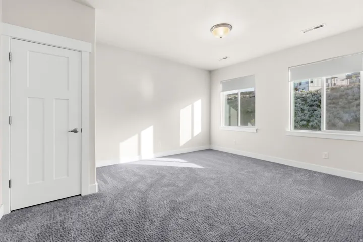 Spacious room featuring patterned gray carpeting, white walls, and two large windows with roller shades