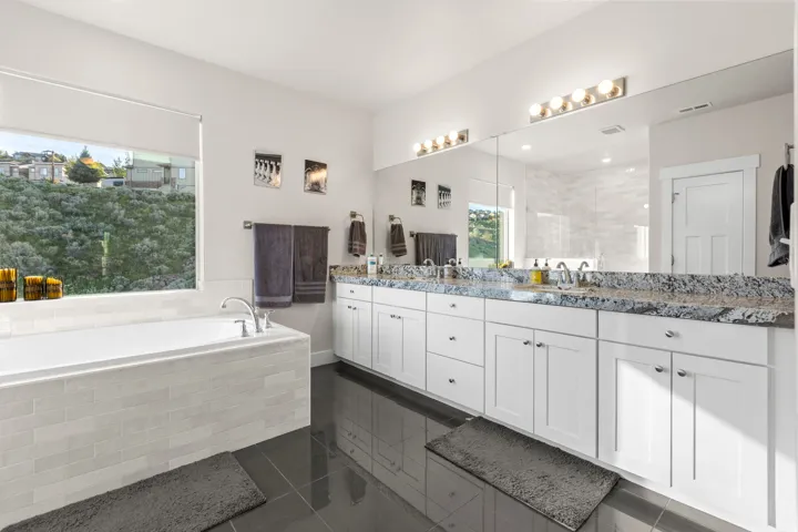 Spacious bathroom featuring a built-in tub with tile surround, a large mirror spanning the vanity, dual white vanities with integrated sinks, and dark tile flooring