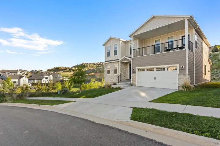 Modern two-story residence featuring a covered second-story balcony with iron railings, a two-car garage with overhead lighting, a concrete driveway, and stone facade accents