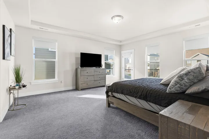 Spacious bedroom featuring light gray carpeting, crisp white walls, multiple windows providing natural light, a tray ceiling, and a glass-paned door opening to an exterior balcony