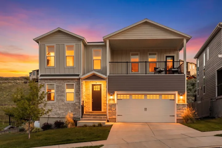 Contemporary facade featuring board and batten siding, stone veneer accents, an upper-level balcony with black railing, a paved driveway, and an integrated garage with transom window detailing