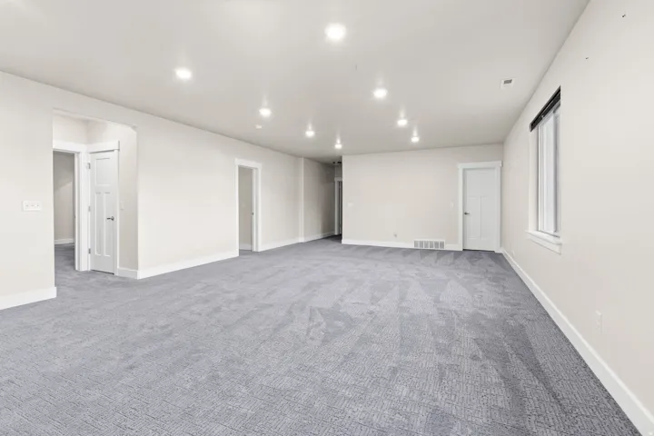 Expansive room featuring light gray patterned carpet, recessed lighting, neutral wall tones, white baseboards, and a window with a dark roller shade
