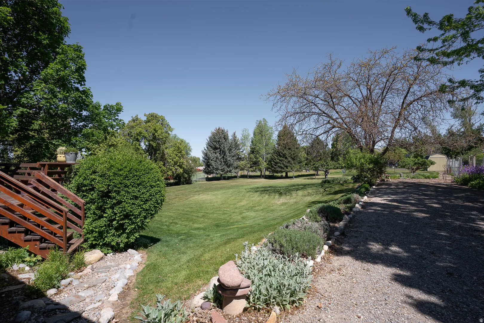Expansive green lawn surrounded by mature trees, featuring a multi-level wood deck with stairs, a gravel driveway, and landscaped garden beds with stone borders