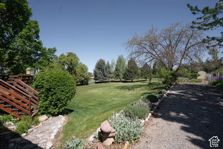 Expansive green lawn surrounded by mature trees, featuring a multi-level wood deck with stairs, a gravel driveway, and landscaped garden beds with stone borders