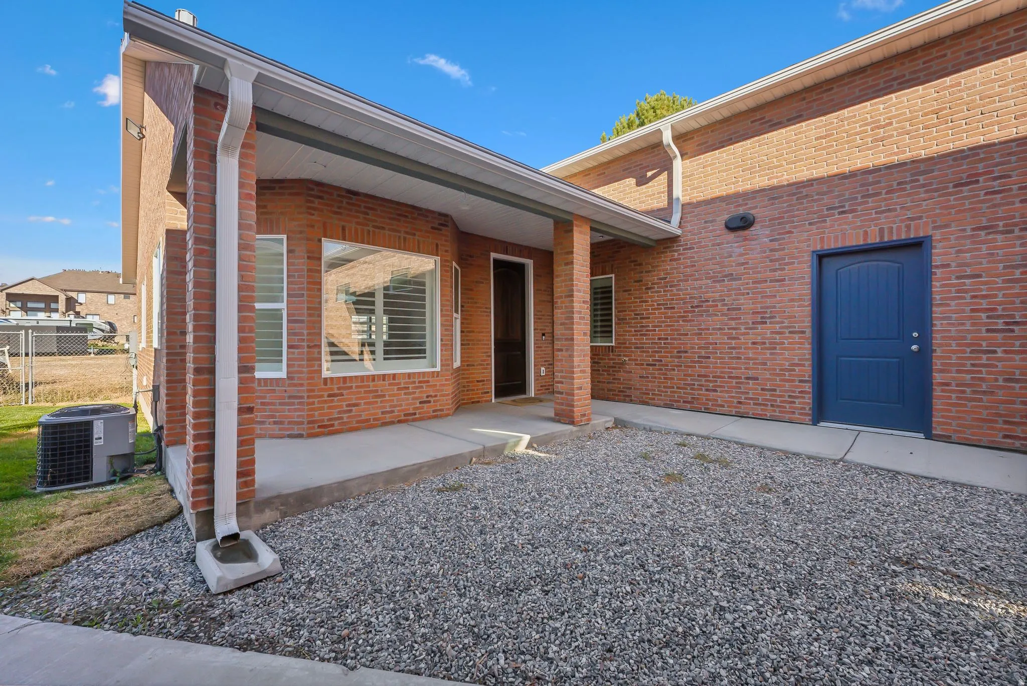 Custom built ADU/Brick exterior featuring a covered porch, solid blue door, bay window with louvered shutters, gravel patio, and concrete walkways
