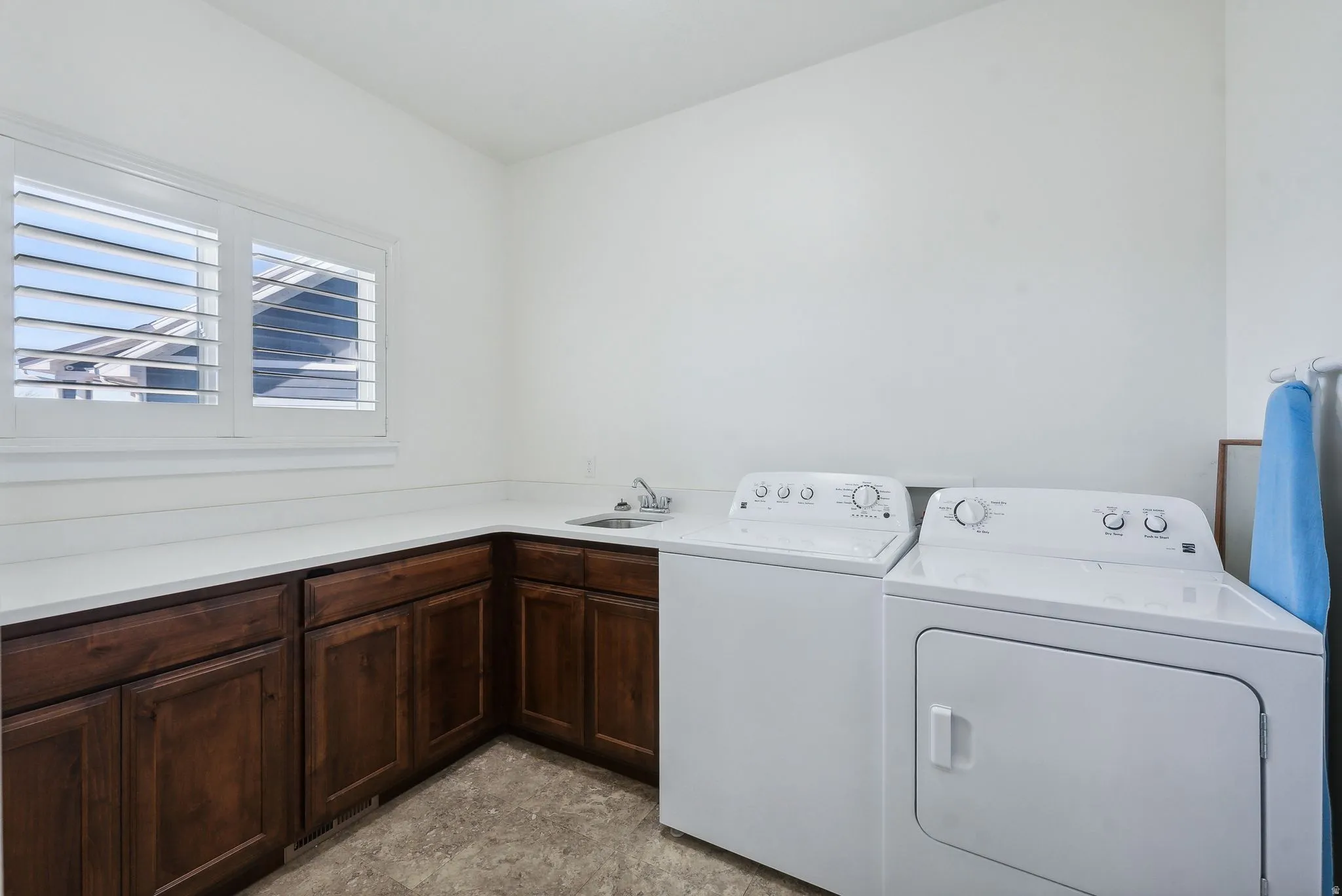 Laundry room featuring L-shaped dark wood cabinetry with white countertops, integrated sink with chrome faucet, and window with white plantation shutters